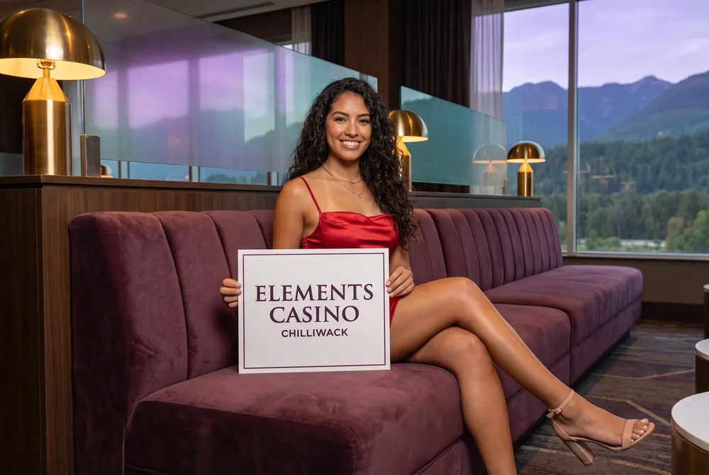 Generated picture of a character created for this guide: a young woman with long dark curls in a red satin mini dress sits on a plum velvet banquette in a lounge at dusk, holding a white card that reads Elements Casino Chilliwack; forested mountains fill the window behind her.